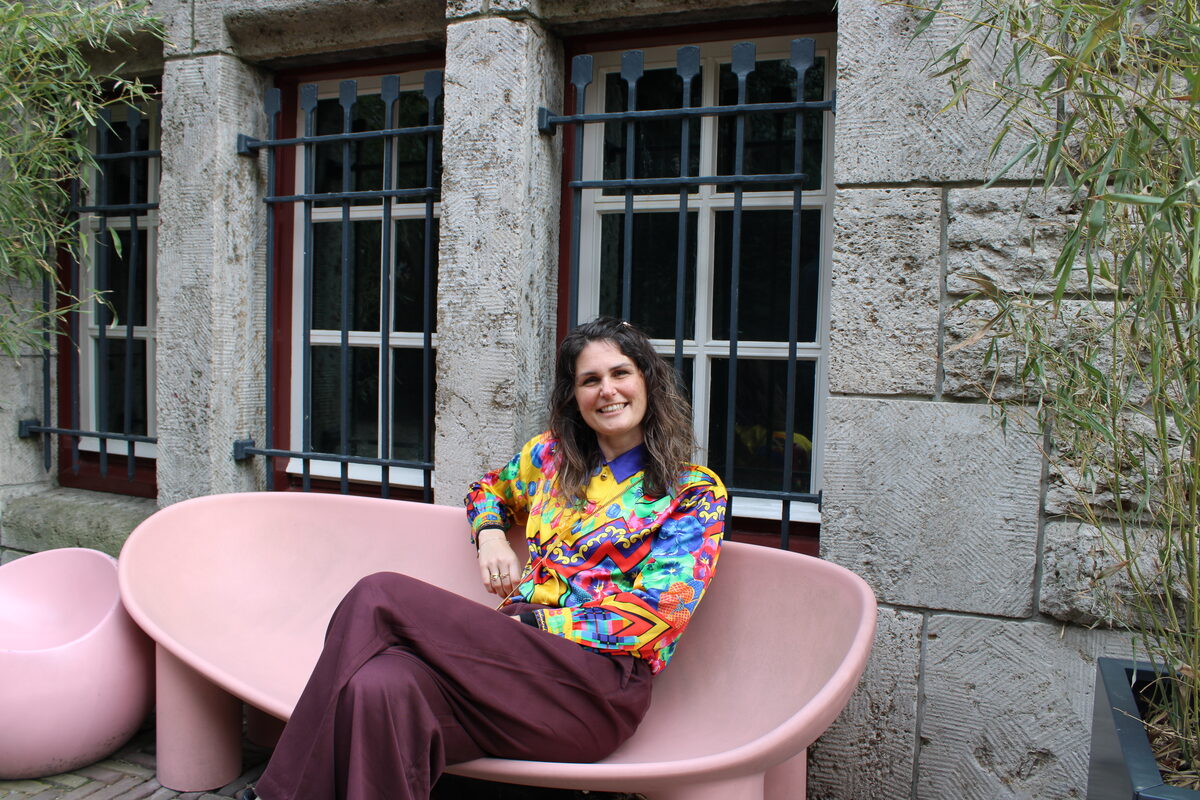 Gabriëlle sitzt in einer farbigen Bluse draußen vor einem Museum auf einer Bank